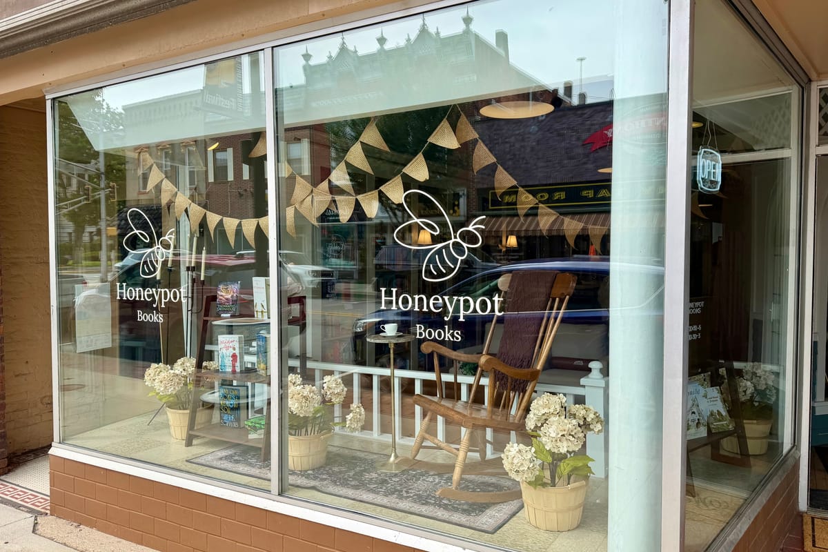 The Honeypot Books storefront: a wide display window with white bee and wordmark decals, burlap bunting, a wooden rocking chair, hydrangeas in baskets, and book displays, with an OPEN sign hanging by the door.